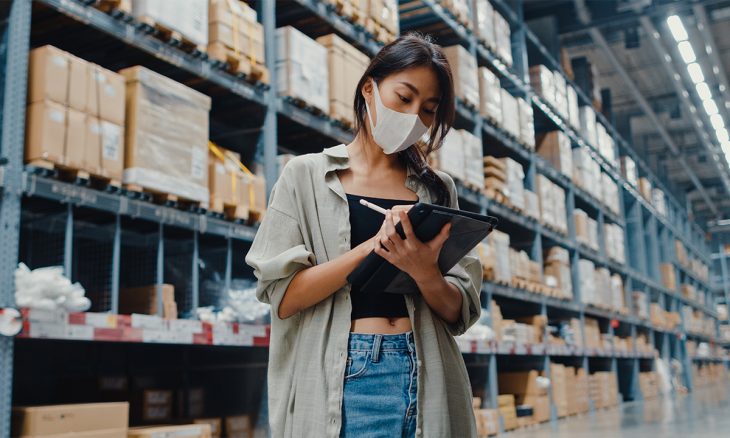 Young Asia businesswoman manager wear face mask looking for goods using digital tablet checking inventory levels stand in retail shopping center. Distribution, Logistics, Packages ready for shipment.